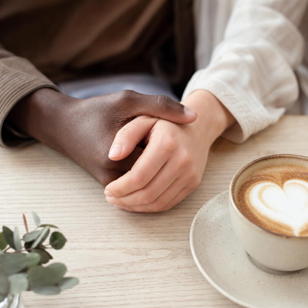 Two hands of an interracial couple holding across a cafe table beside a cappuccino with a heart in the foam